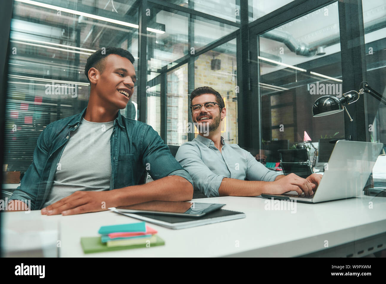 Atmosphère positive. Deux jeunes et beaux hommes à parler les uns avec les autres et souriant tout en travaillant dans un bureau moderne. Les gens d'affaires. Lieu de travail. Le travail d'équipe Banque D'Images