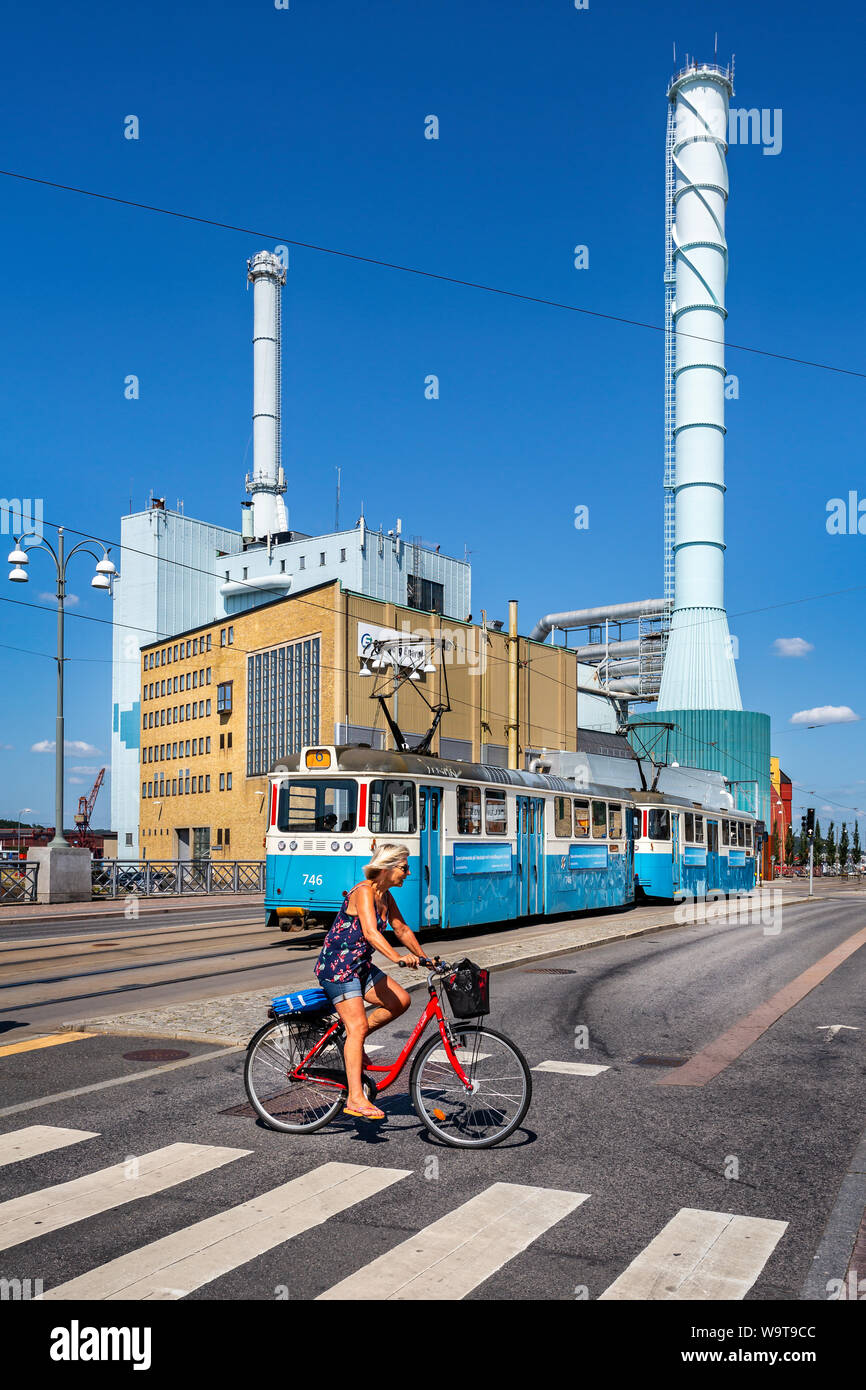 Dame cycliste sur route passage passage clouté avec le tram et les cheminées des centrales électriques à l'arrière-plan à Göteborg, en Suède le 26 juillet 2019 Banque D'Images