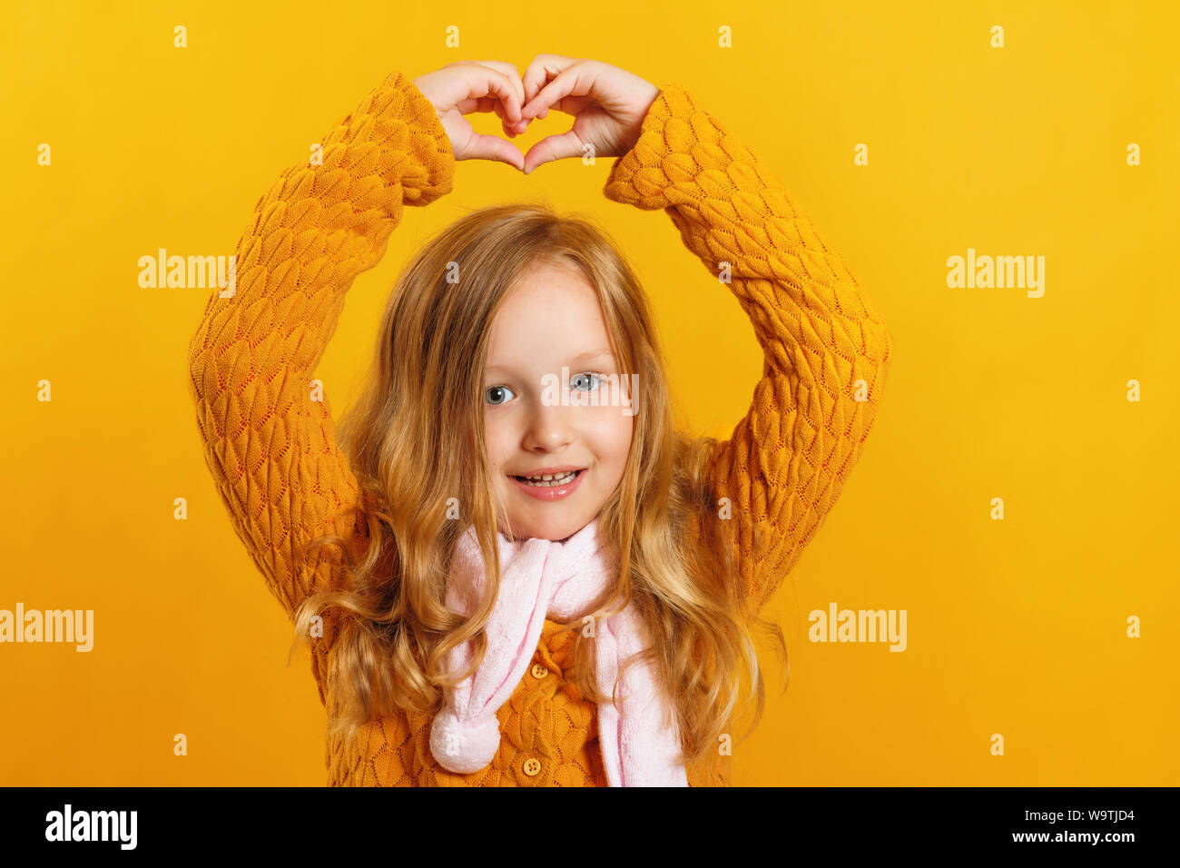 Petite fille joyeuse dans un pull et écharpe, montre un coeur au-dessus de  sa tête avec ses mains, regardant la caméra sur un fond jaune Photo Stock -  Alamy
