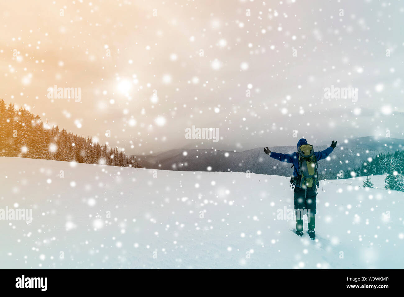 Vue arrière de l'randonneur dans des vêtements chauds avec sac à dos debout avec les bras levés sur la compensation couverte de neige sur la montagne de la forêt d'épinettes et ciel nuageux Banque D'Images