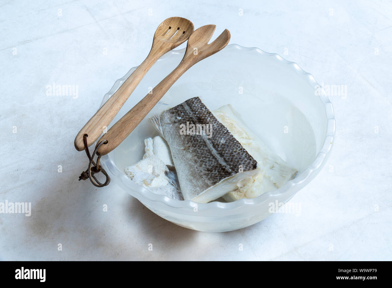 La morue salée et séchée (Gadus morhua) dans un bol en plastique rempli d'eau à dessaler et être cuits et cuillère en bois sur fond blanc. Alimentation saine. Banque D'Images