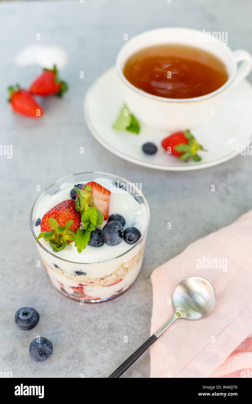 Gruau avec du yogourt avec des fraises et des bleuets avec cuillère et une tasse de thé sur fond gris avec une serviette rose et cuillère. Concept de pique-nique avec Banque D'Images Gruau avec du yogourt avec des fraises et des bleuets avec cuillère et une tasse de thé sur fond gris avec une serviette rose et cuillère. Concept de pique-nique avec Banque D'Images
