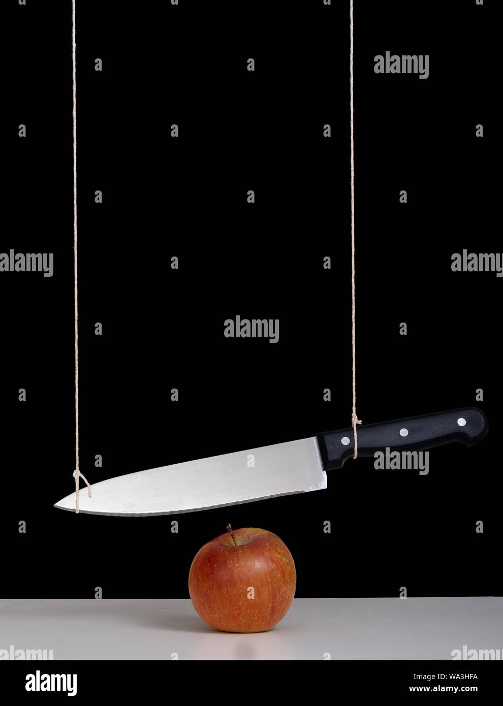 Épée de Damoclès menace, notion de risque, métaphore - grand couteau attaché et suspendu au-dessus d'Apple. Nature morte avec fond noir. Banque D'Images