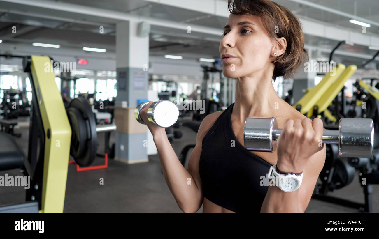 Portrait de femme mature athlétique fait des représentants de l'exercice pour les biceps avec haltères en mains dans une salle de sport. Elle est levée haltères courtes, copier l'espace. La formation et le sport concept. Banque D'Images