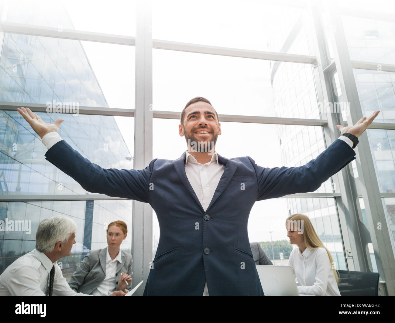 Joyeux jeune businessman standing with arms raised smiling , équipe de ses collègues assis à la table dans l'arrière-plan Banque D'Images