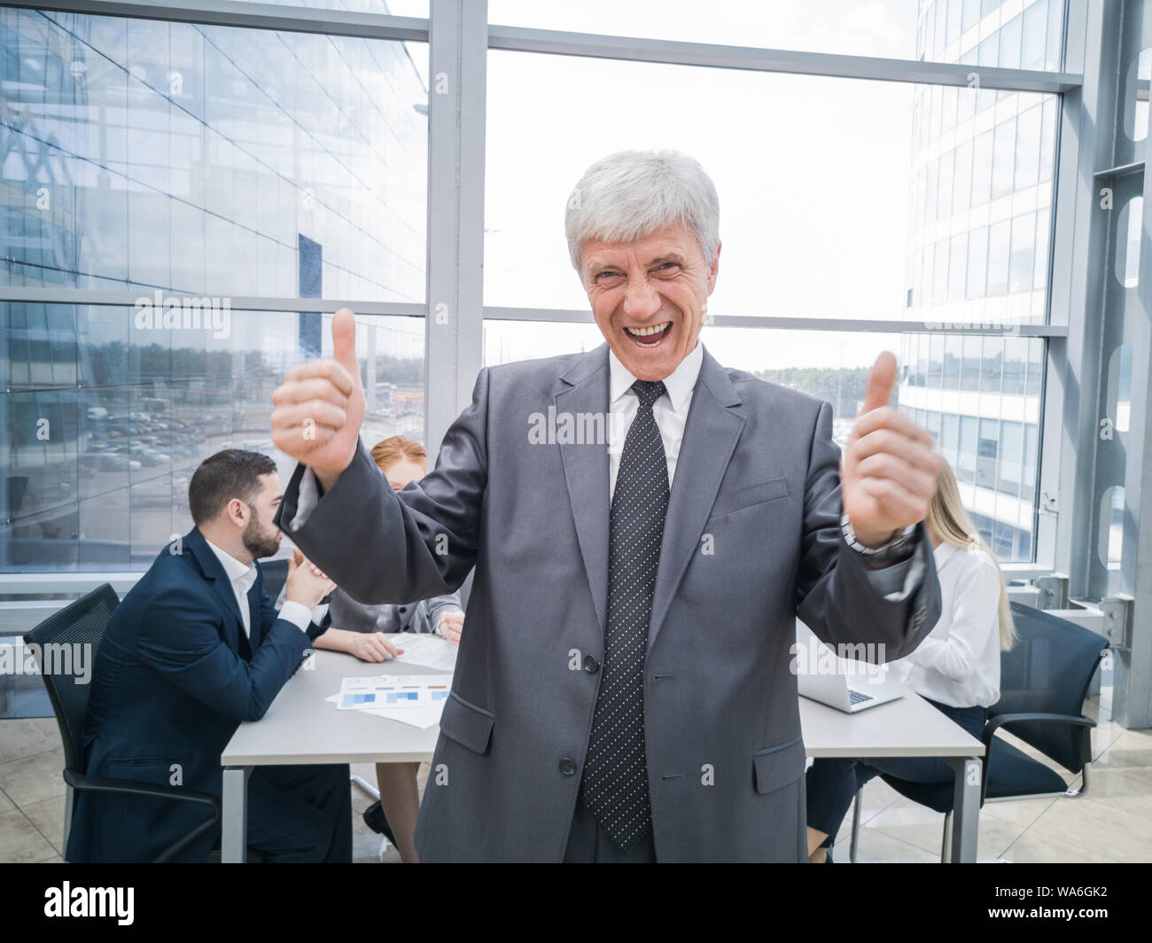 Confiant mature businessman showing thumb up and smiling , équipe de ses collègues assis à la table dans l'arrière-plan Banque D'Images
