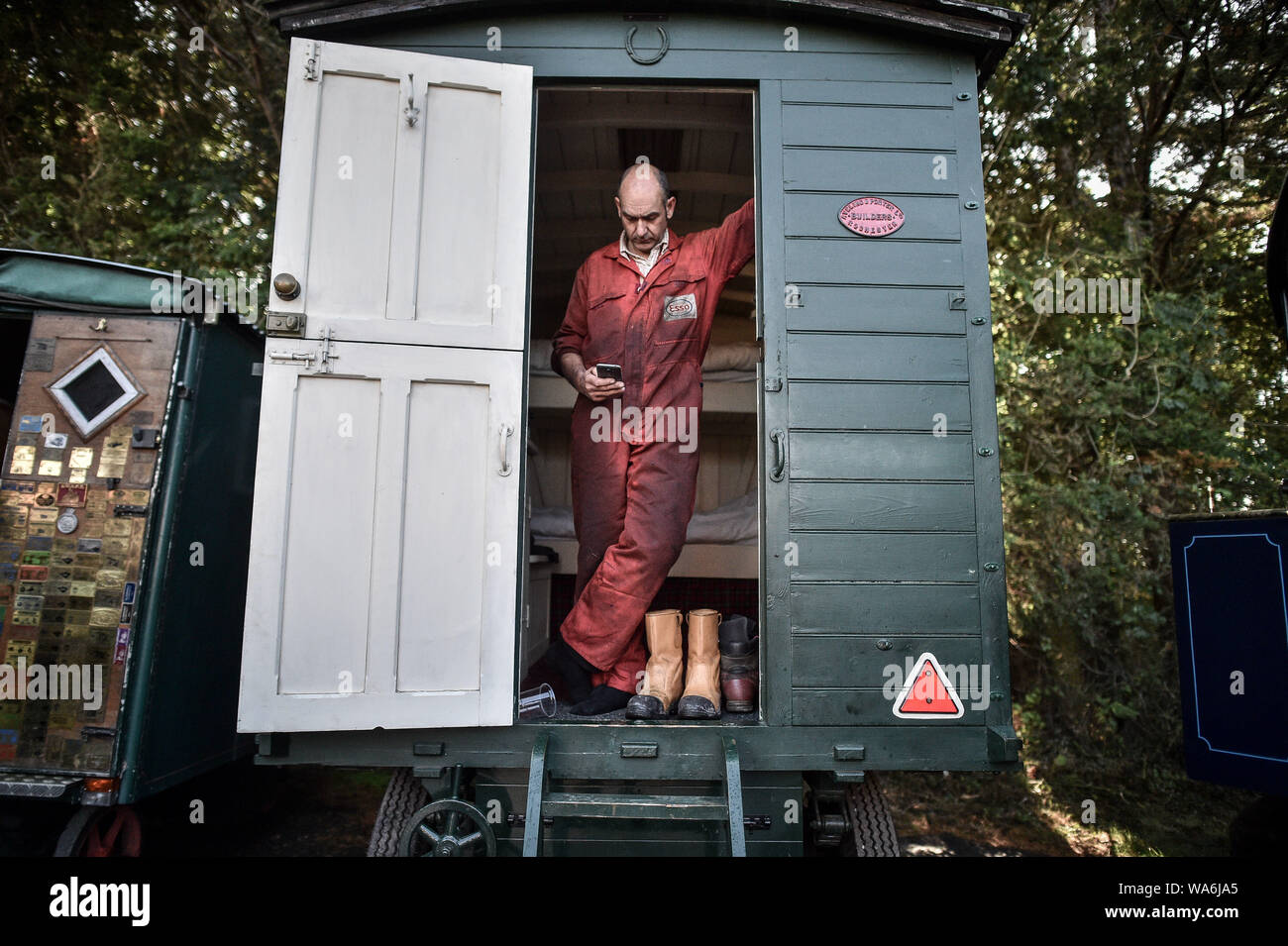 Un homme en salopette huileux vérifie son téléphone de sa caravane à Drusilla's Inn, près de Horton, Dorset, comme des dizaines de véhicules à vapeur se préparent à faire le voyage jusqu'à la grande foire de vapeur Dorset, où ils seront rejoints par des milliers de passionnés de célébrer la puissance de la vapeur à partir de 22 Août sur le week-end férié. Banque D'Images