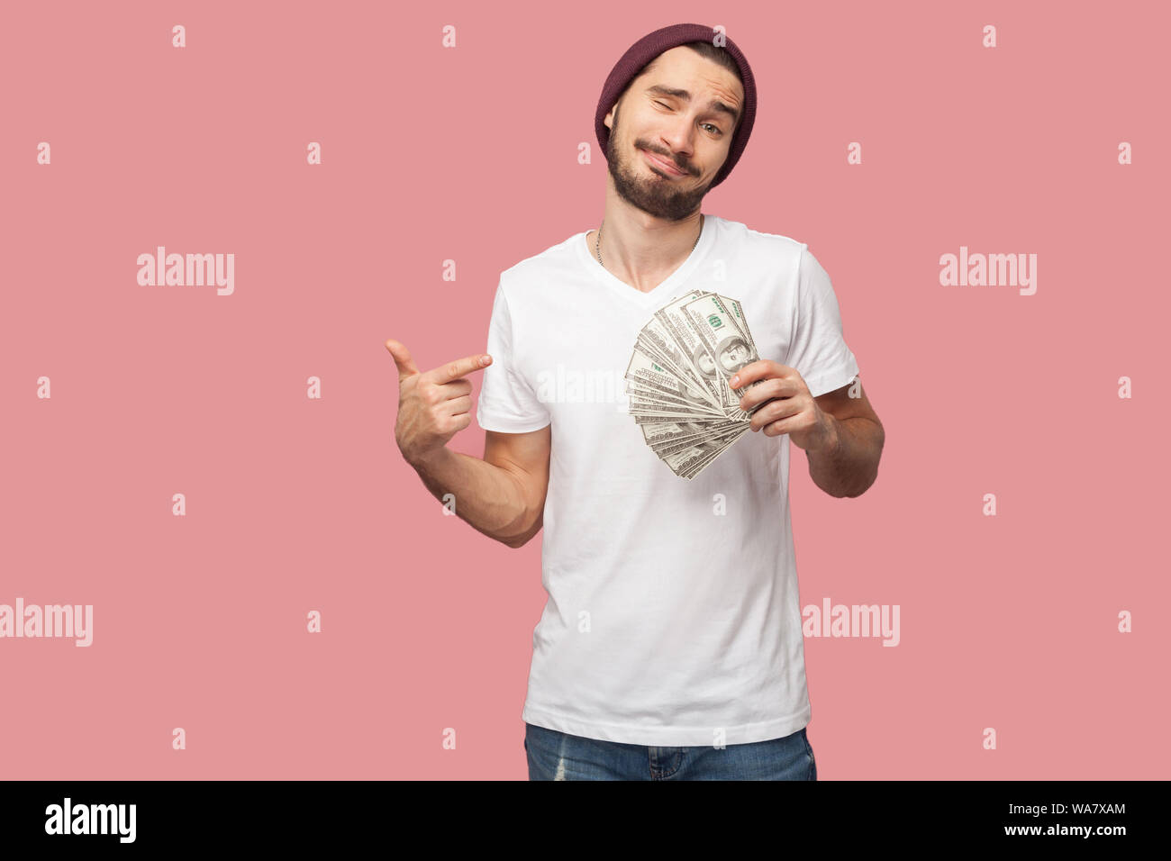 Je suis homme riche. Beau jeune homme hipster barbu en chemise blanche et chapeau décontracté, le socle, le holding fan de dollars de l'argent et montre de l'index de l'argent. Je Banque D'Images