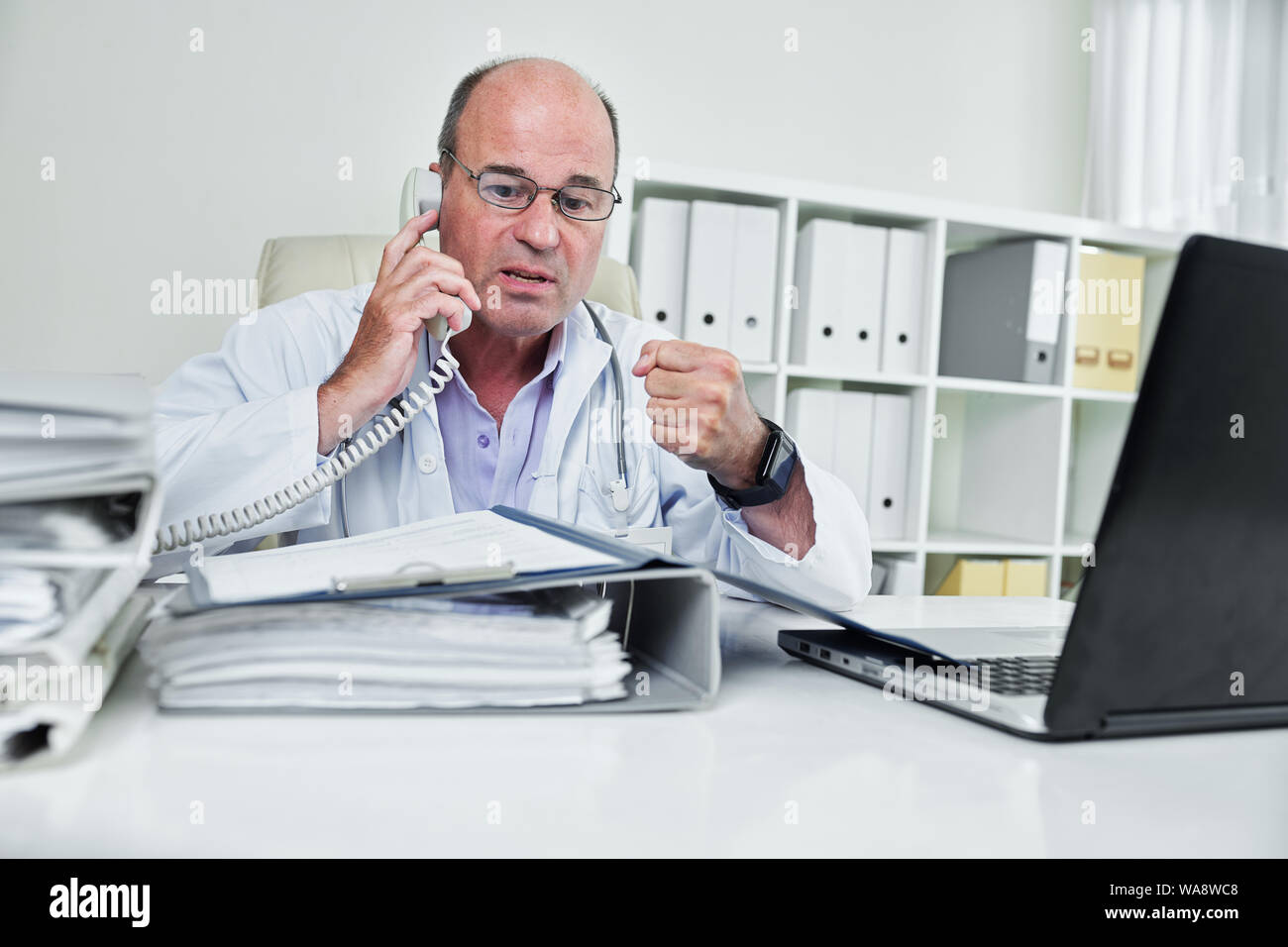Mature en colère médecin chef à la recherche de document dans le dossier ouvert sur sa table et chouting au récepteur du téléphone Banque D'Images