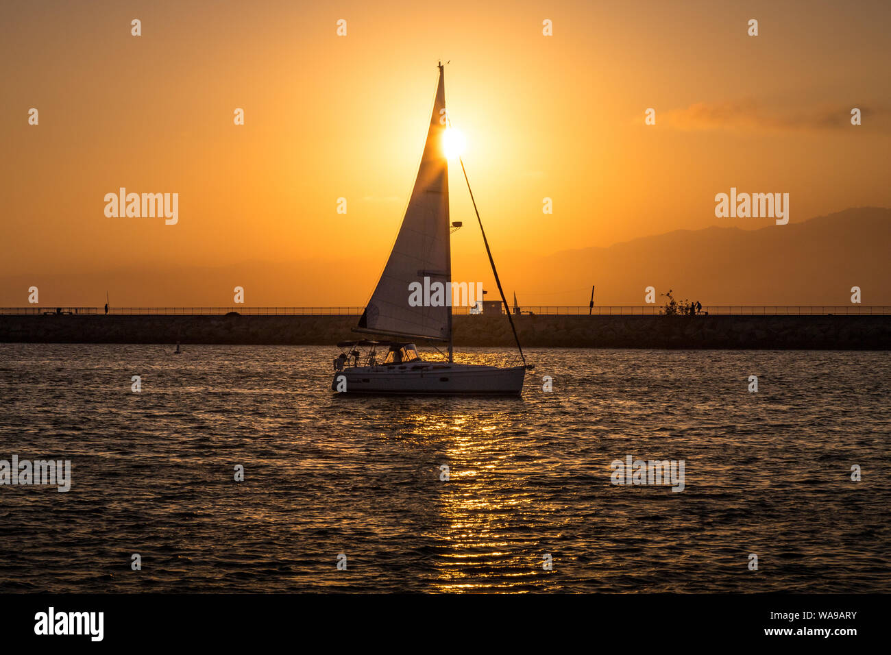 Voilier naviguant dans Marina del Rey, Californie au coucher du soleil avec des nuages dans le ciel et le Santa Monica montagnes en arrière-plan Banque D'Images