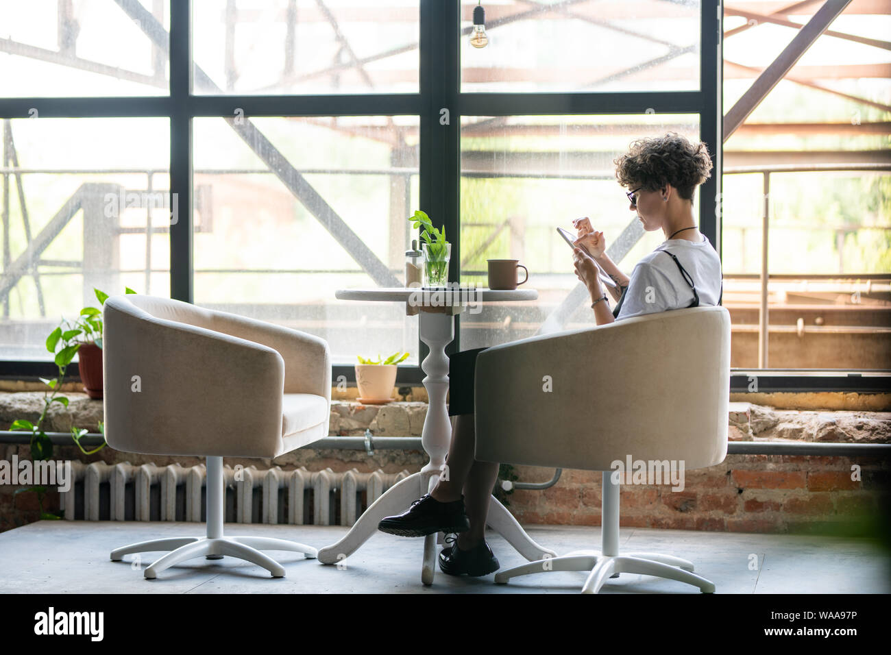 Jeune femme avec gadget mobile sitting in armchair par fenêtre dans studio Banque D'Images