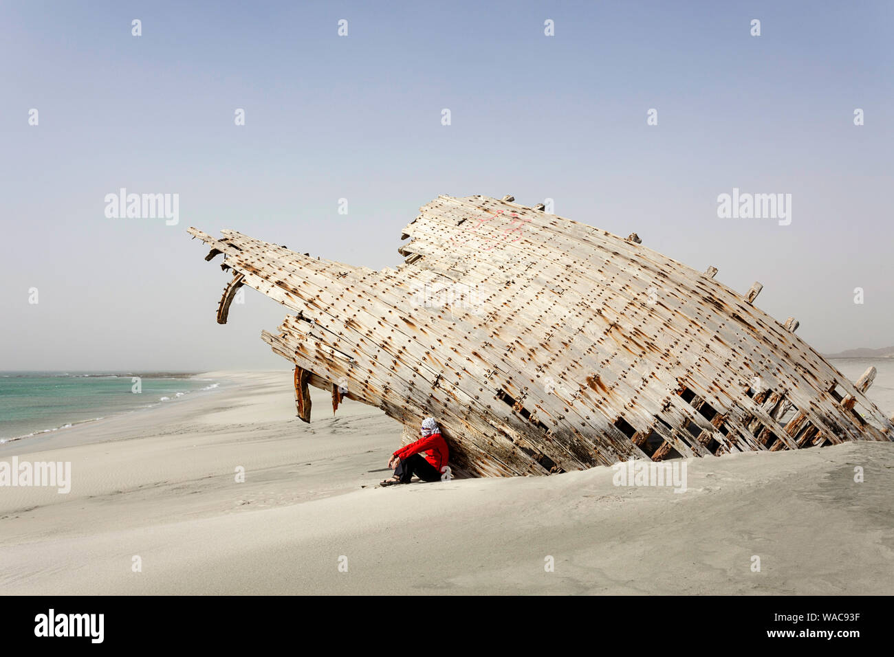 Homme portant un foulard assis sous le bateau 'Perdu', l'épave d'un boutre sur une plage de sable blanc sur la côte est de l'île de Masirah, Oman Banque D'Images