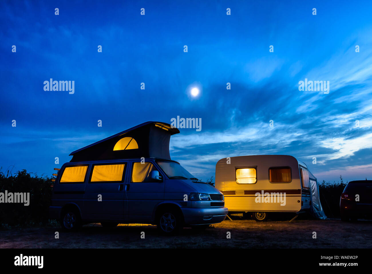 Un petit camping-van garé à côté d'une caravane vintage dans un camping,  tous les deux allumés de l'intérieur à la tombée de la nuit avec la lune  brillants dans un ciel d'orage