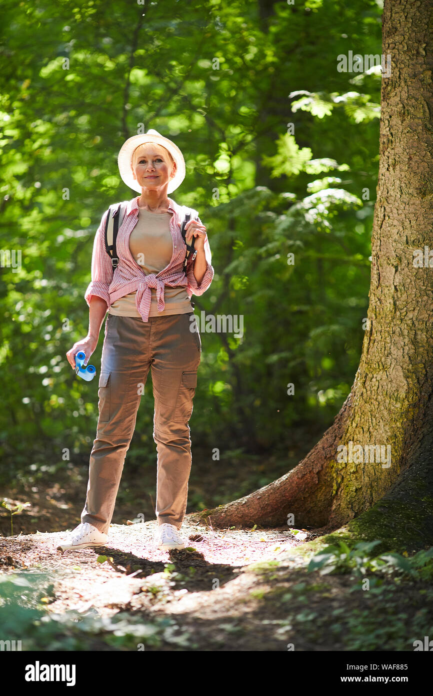 Portrait of mature woman in hat et avec sac à dos à la caméra à tout en se tenant dans la forêt durant son voyage aventure Banque D'Images