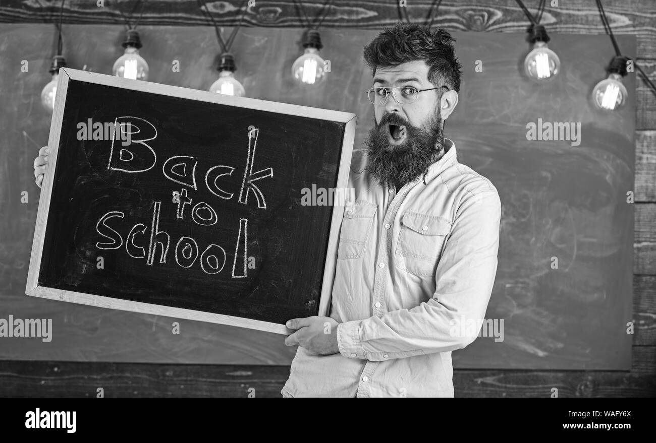 Homme avec barbe et moustache sur le visage choqué accueille des étudiants, tableau sur l'arrière-plan. L'éducation et l'étude de concept. Dans l'enseignant détient le titre de lunettes avec tableau noir retour à l'école. Banque D'Images