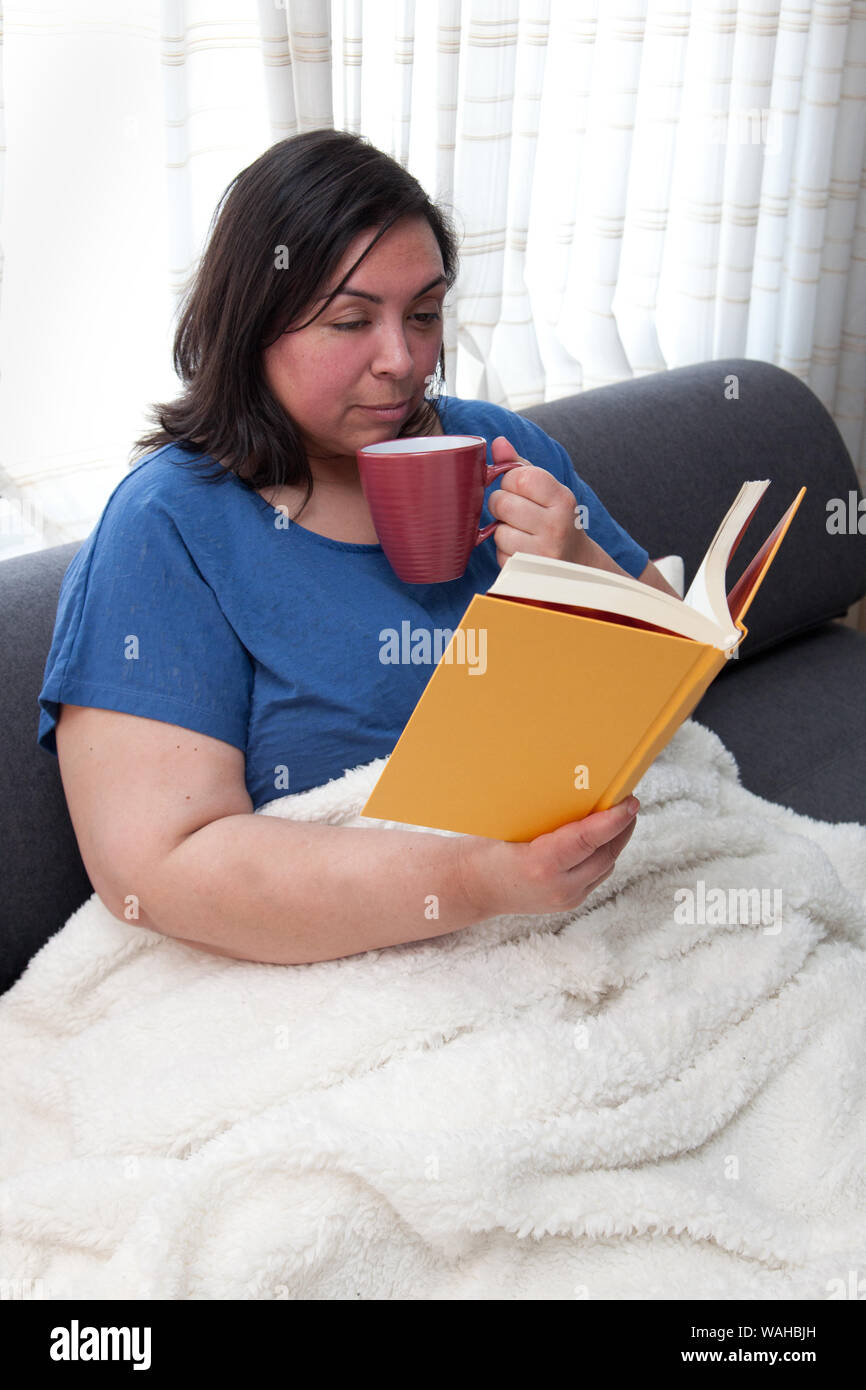Du vrai latina woman cooling off sa tasse de boisson chaude pendant la lecture d'un roman à la couverture rigide Banque D'Images