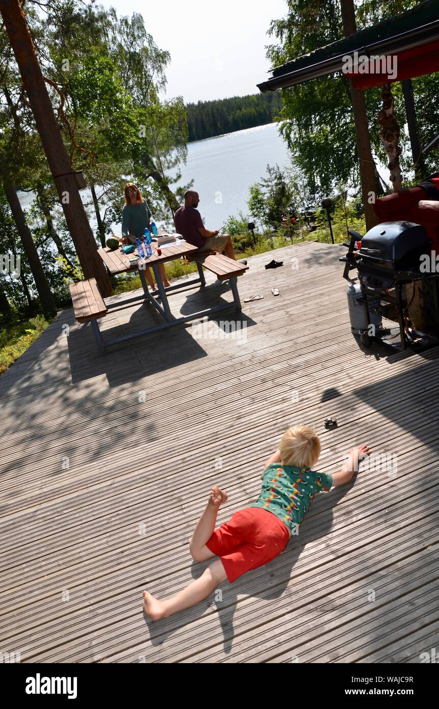 Jeune garçon blond jouant sur decking en bois à l'extérieur de log cabin, parents en arrière-plan, le lac de Sarkjarvi, Finlande. Banque D'Images