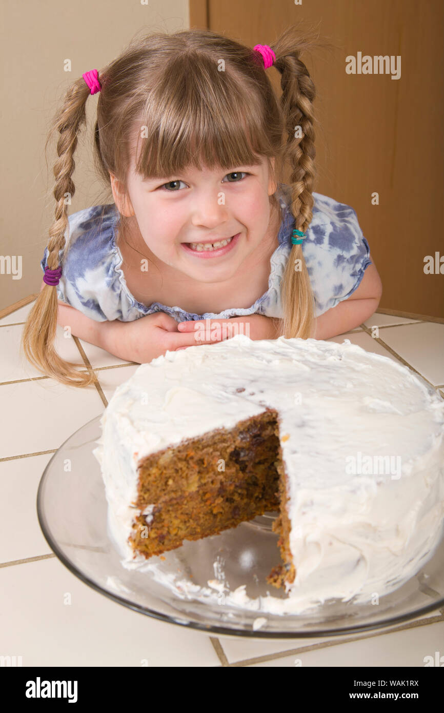 Jeune fille en face d'un gâteau aux carottes avec une tranche coupée, avec glaçage à la crème, reposant sur un verre cake stand. (MR, communication) Banque D'Images
