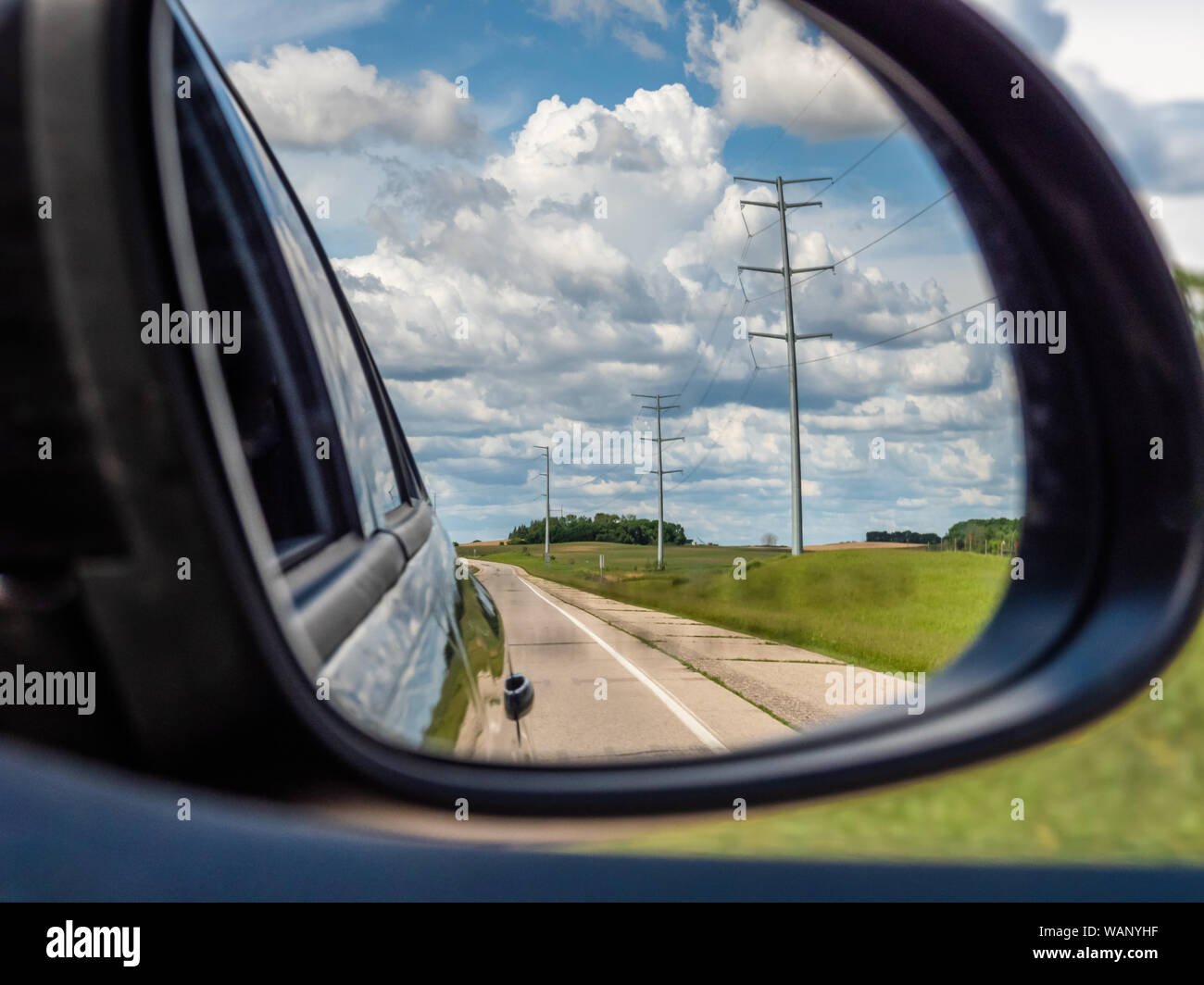 Voir retour derrière de la campagne agricole en milieu rural avec les nuages et les lignes électriques le long d'une route encadrée dans le rétroviseur latéral d'une voiture en mouvement. Banque D'Images