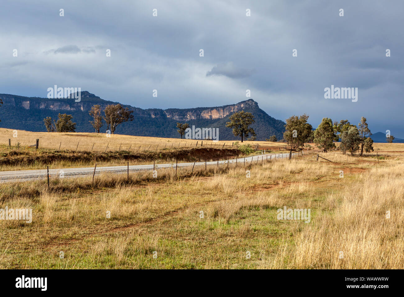 Vide chemin rural traversant les plaines herbeuses dans la vallée Capertee, NSW, Australie, avec des crêtes de grès et le bush naturel dans l'arrière-plan Banque D'Images