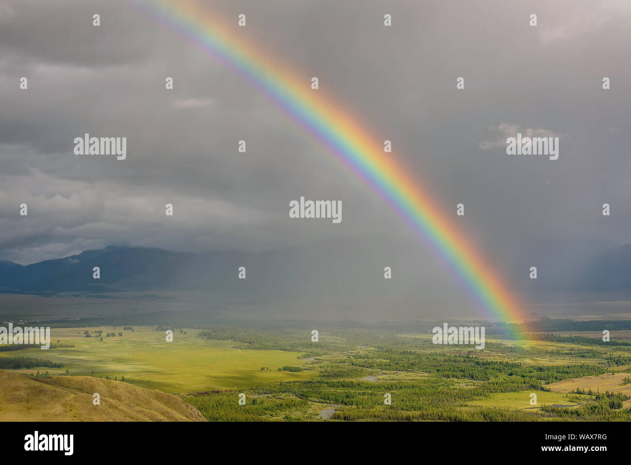 Arc-en-ciel de couleurs vives incroyable sur les montagnes, une vallée avec une rivière sinueuse et la forêt contre un ciel d'orage avec des nuages et de la pluie. L'Altaï, en Russie. Banque D'Images