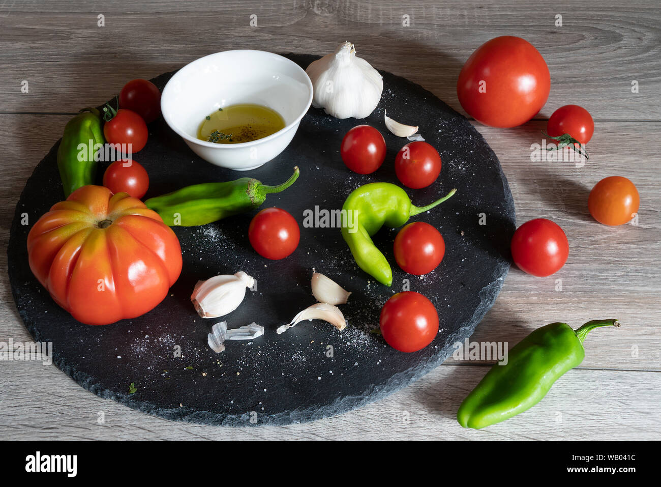 Quelques tomates, gousses d'ail et un bol avec de l'huile d'olive noire sur une plaque en pierre et une table en bois Banque D'Images