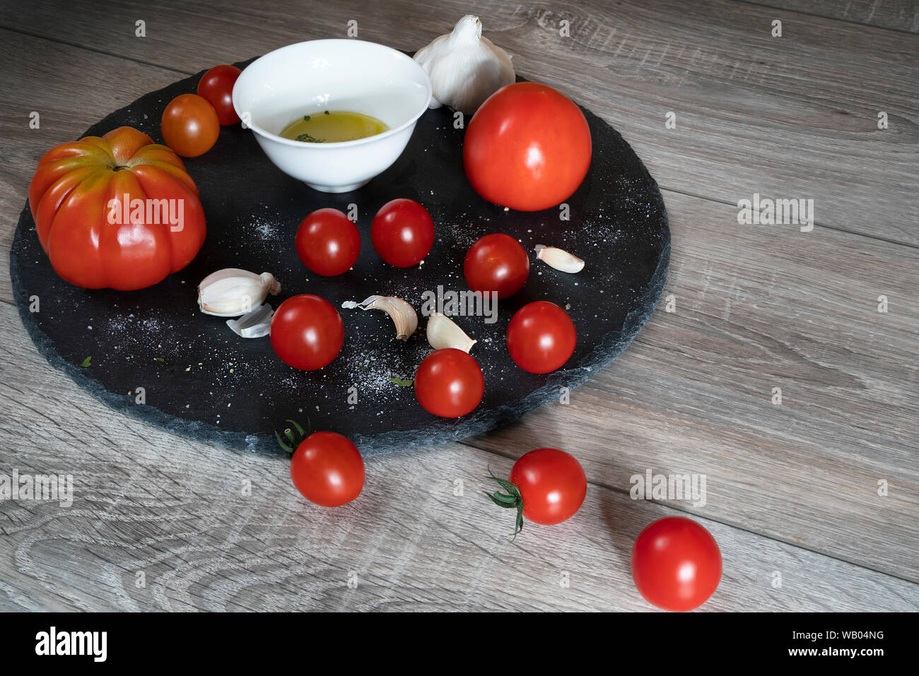 Quelques tomates, gousses d'ail et un bol avec de l'huile d'olive noire sur une plaque en pierre et une table en bois Banque D'Images