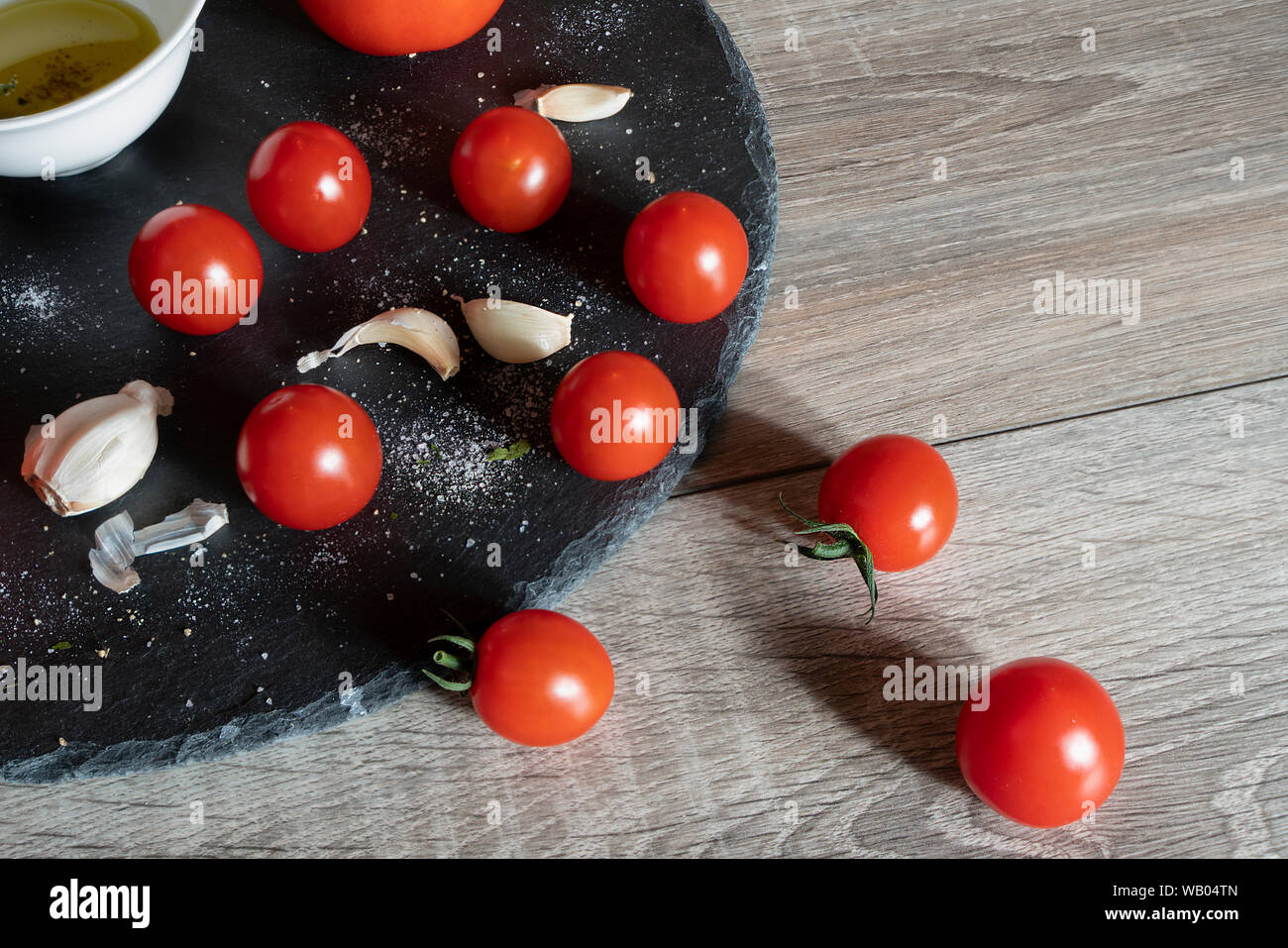 Quelques tomates, gousses d'ail et un bol avec de l'huile d'olive noire sur une plaque en pierre et une table en bois Banque D'Images