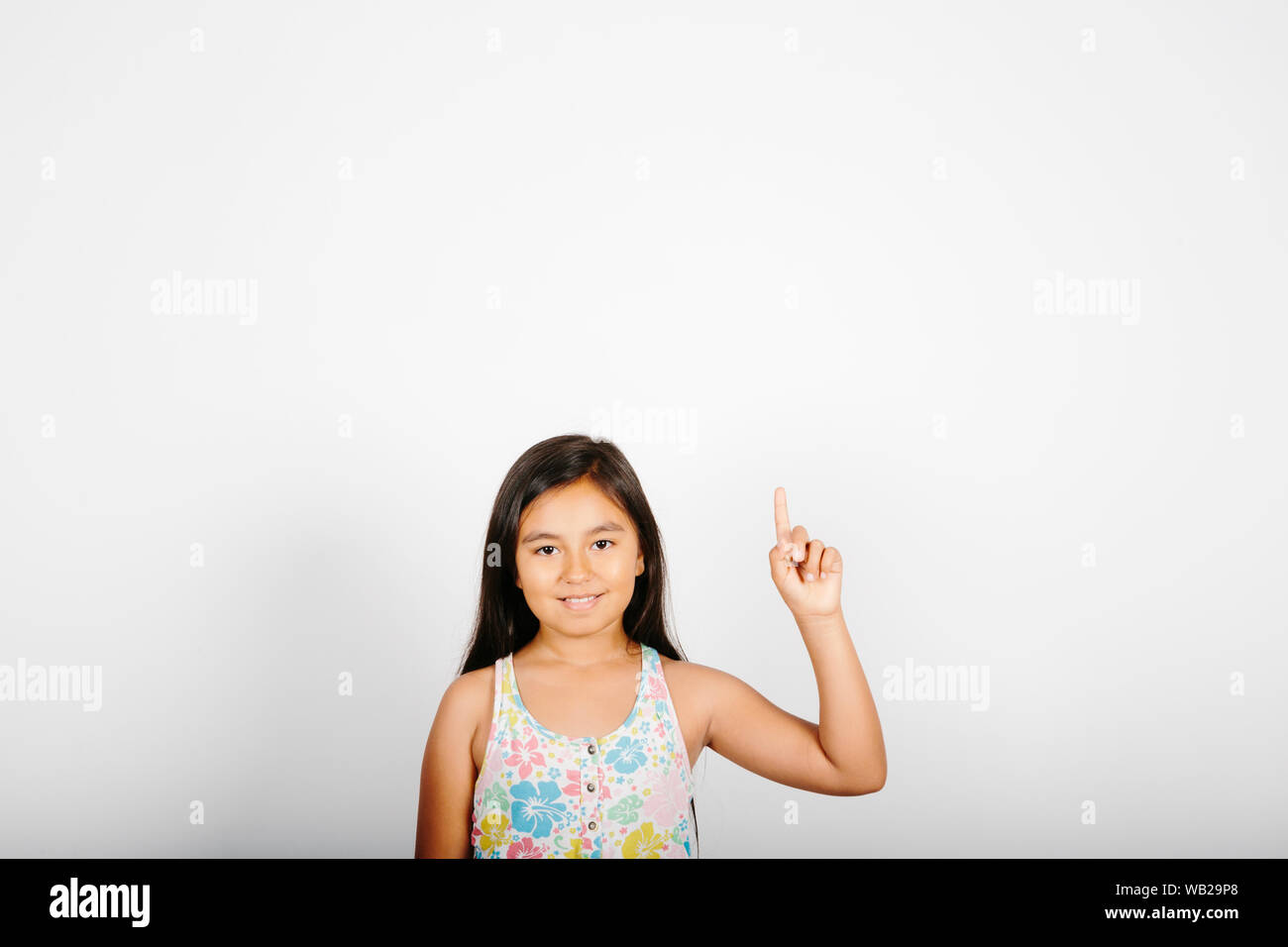 Une fillette de 9 ans est orientée vers le haut. Elle est debout devant un mur blanc. Elle est souriante. Elle a de longs cheveux noirs. Elle a les caractéristiques d'origine hispanique. Elle a wea Banque D'Images