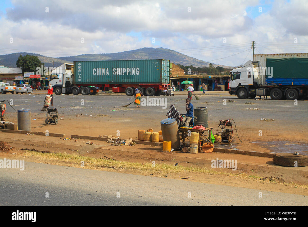 Conteneur d'expédition chinois sur camion transporté par la route au Kenya. Les gens balayant la route poussiéreuse et vendant le commerce sur le côté de la route. Afrique Banque D'Images