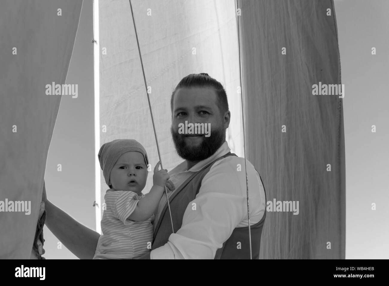 Homme barbu tenant un bébé dans ses bras contre le fond de ciel et navire, monochrome Banque D'Images