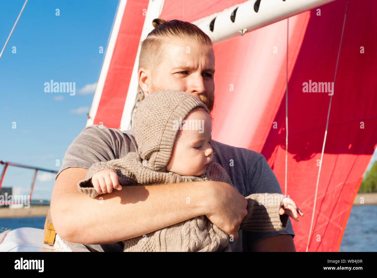 Homme barbu tenant un bébé dans ses bras sur le fond bleu du ciel et un mât de navire à voiles rouge Banque D'Images