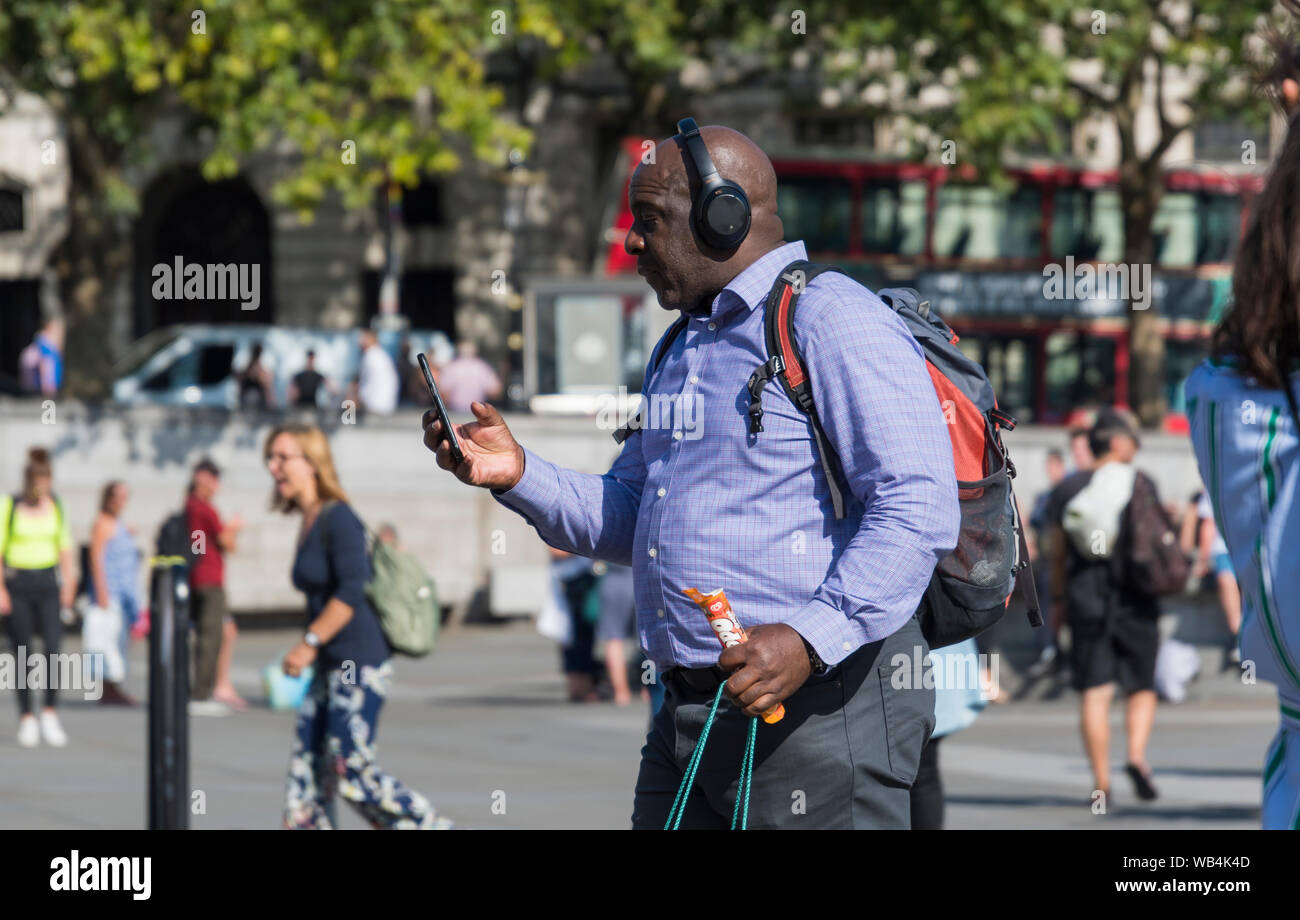Bald Middle aged black man walking in Summer portant des écouteurs et grande à l'aide d'un smartphone dans le centre de Londres, Angleterre, Royaume-Uni. Banque D'Images