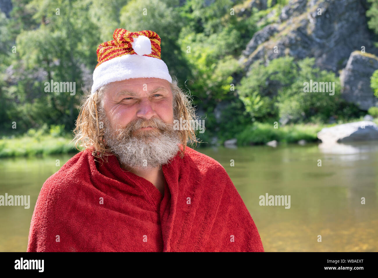 Vieil homme charismatique avec une barbe dans un Santa Claus Hat et Red Coat smiling. Dans le contexte d'un paysage d'été, rivière, montagne, forêt Banque D'Images