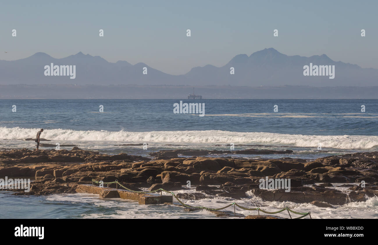Mossel Bay, Afrique du Sud - un vacancier explore les rochers à la Punt beach avec une plate-forme pétrolière dans la baie à l'arrière-plan Banque D'Images