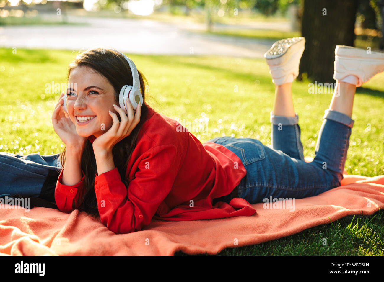 Image de laughing girl student wearing red jacket à l'écoute de la musique avec écouteurs en lying on blanket in green park Banque D'Images