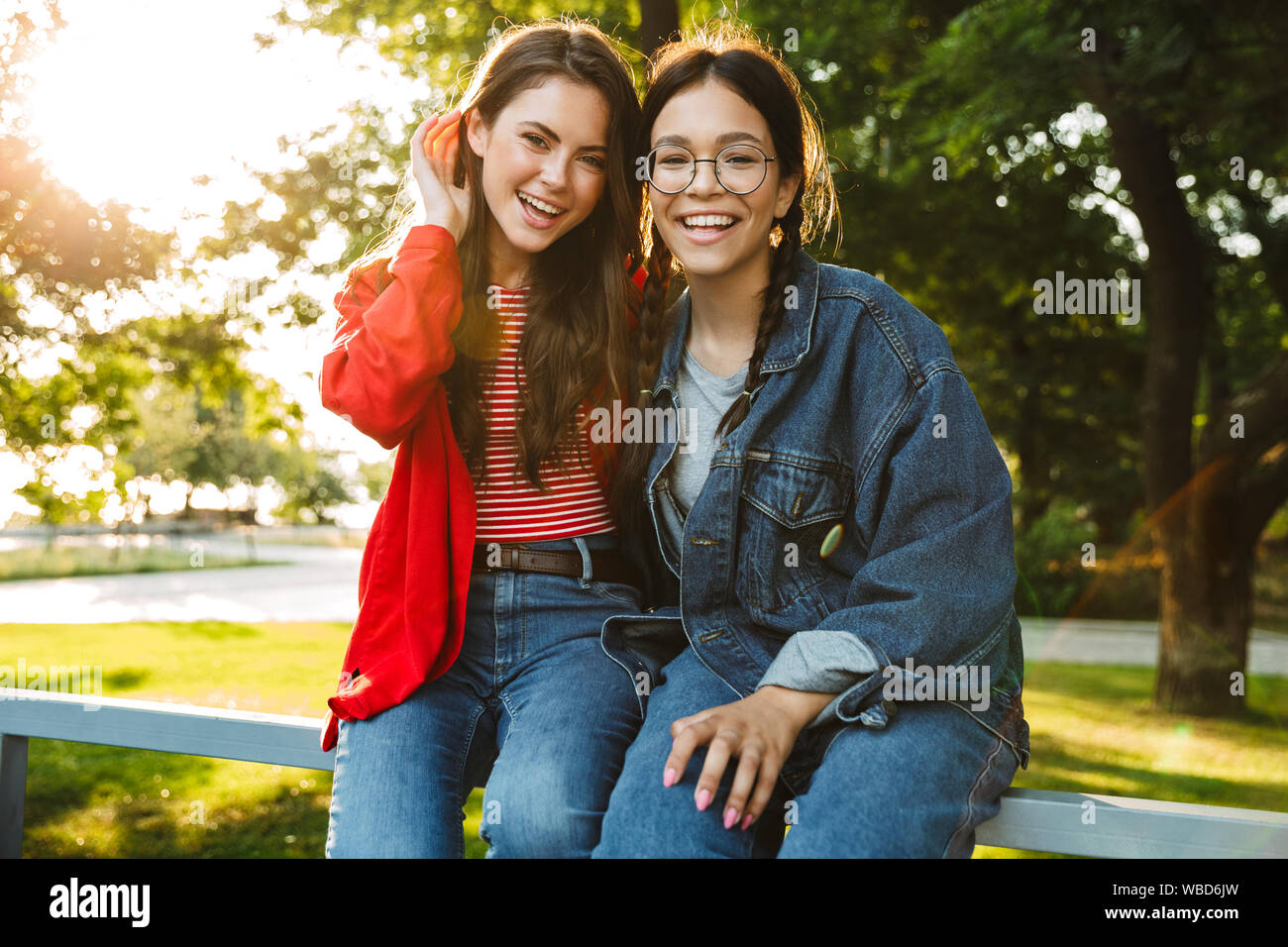 Image de deux élèves filles gaie et souriante hugging while sitting on railing in green park Banque D'Images