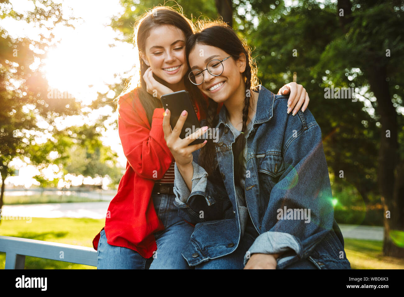 Image de deux étudiants smiling girls using cellphone et étreindre tout en restant assis sur main courante en green park Banque D'Images