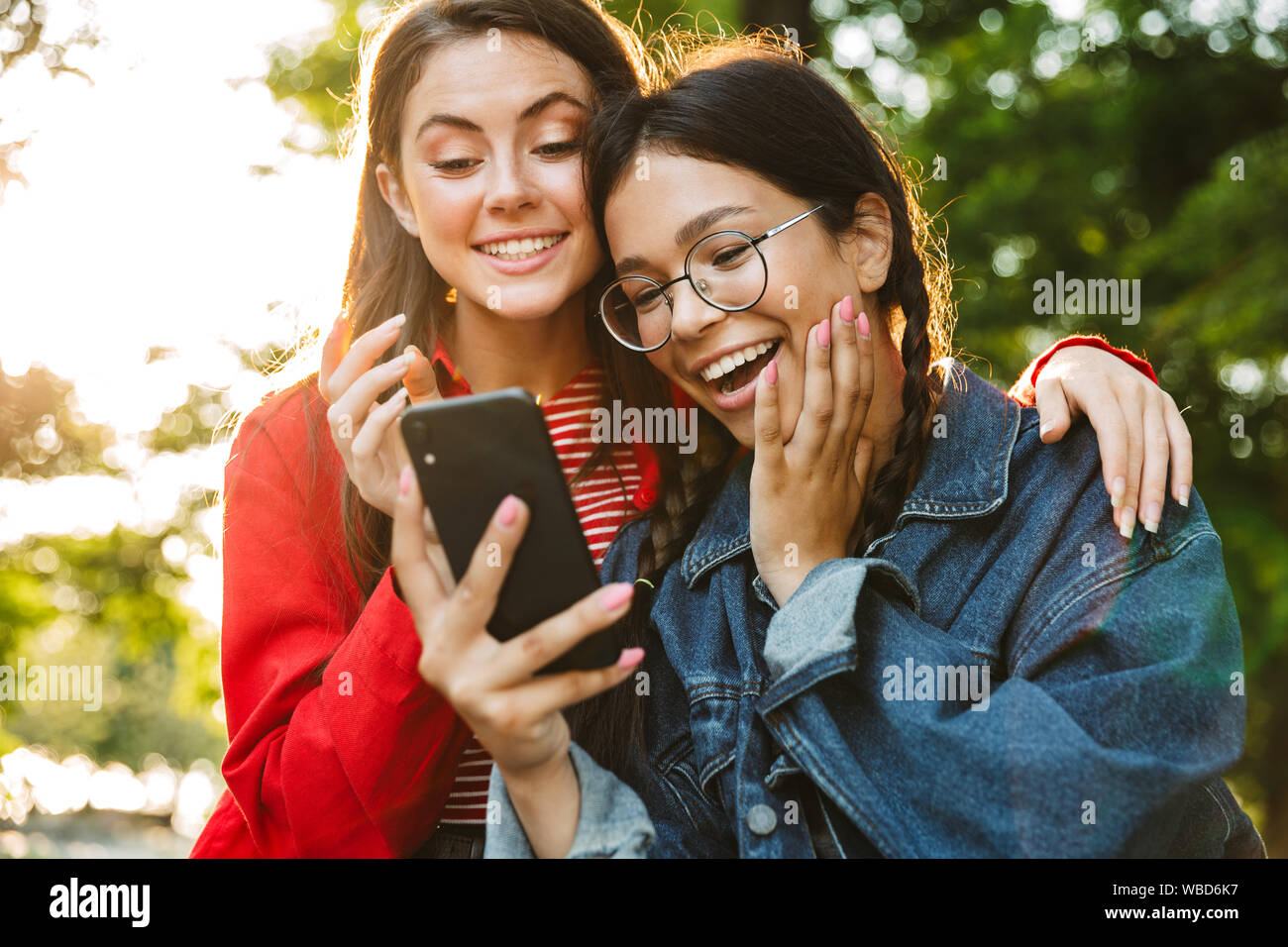 Image de deux élèves filles excité à l'aide de téléphone cellulaire et d'étreindre tout en restant assis sur main courante en green park Banque D'Images