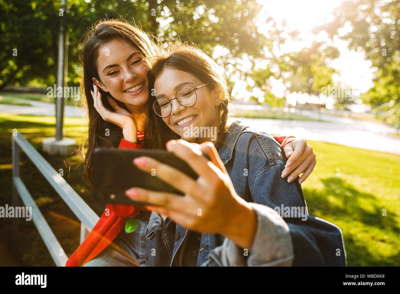 Image de deux jolies filles élèves prenant photo selfies sur téléphone cellulaire et souriant tout en restant assis sur main courante en green park Banque D'Images