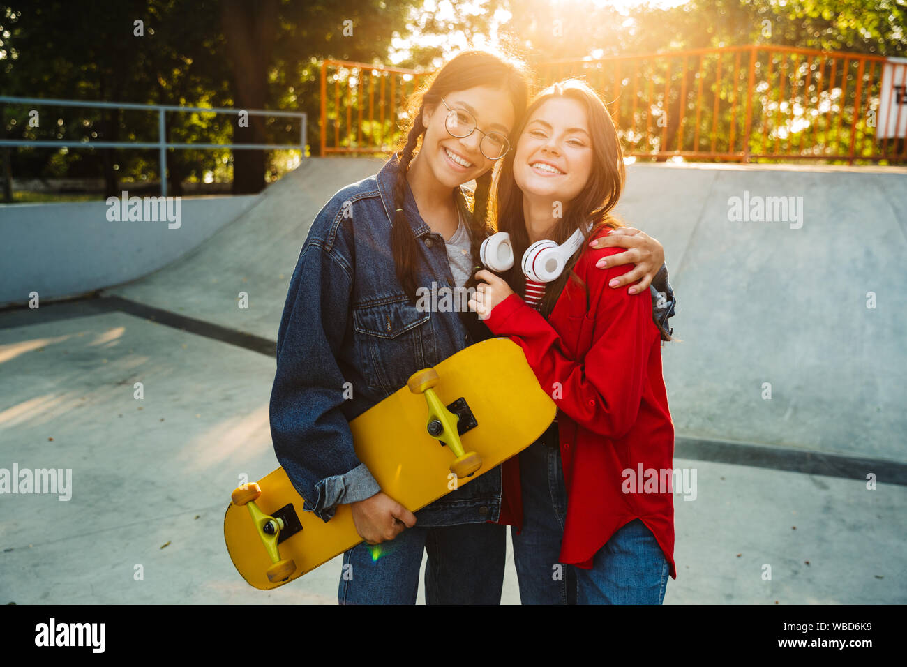 Image de deux filles joyeux habillés en vêtements de denim en souriant et serrant ensemble tout en maintenant en skateboard skate park Banque D'Images