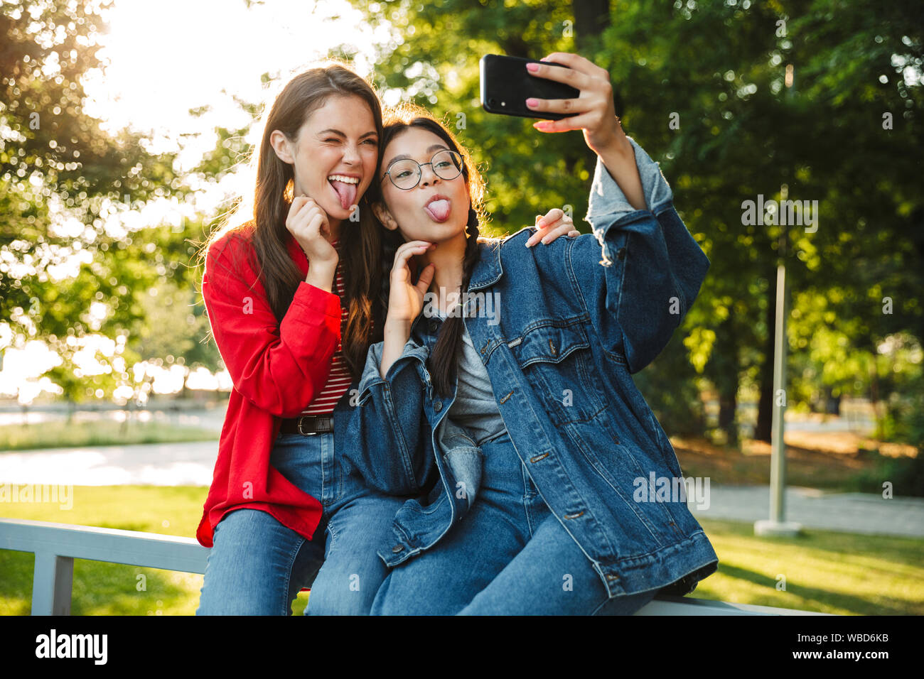 Image de deux étudiants en funny girls photo selfies sur téléphone cellulaire en langues qui sort tout en restant assis sur main courante en green park Banque D'Images