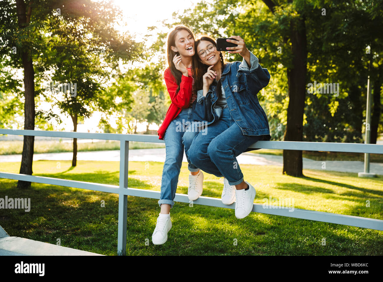 Image de deux élèves filles heureux de prendre photo sur selfies cellulaire et un sourire tout en restant assis sur main courante en green park Banque D'Images