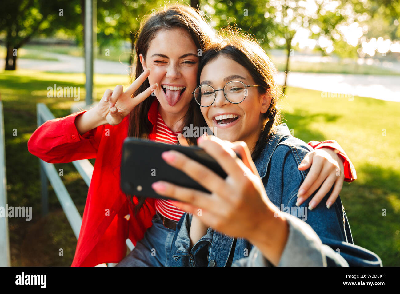 Image de deux étudiants en funny girls photo selfies on cellphone and gesturing while sitting on chanter la paix railing in green park Banque D'Images