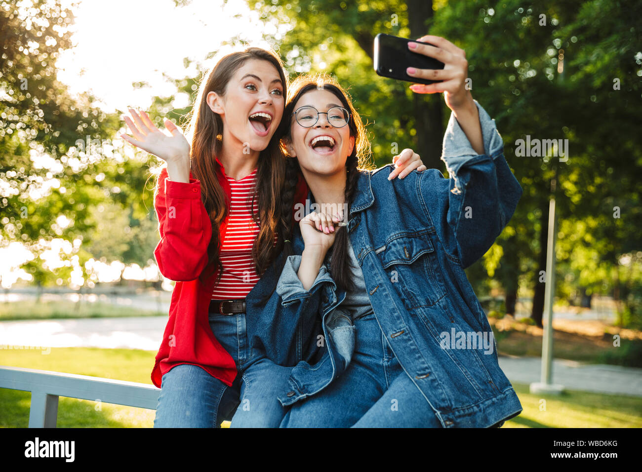 Image de deux élèves filles étonné prendre photo sur téléphone portable et selfies hugging tout en restant assis sur main courante en green park Banque D'Images