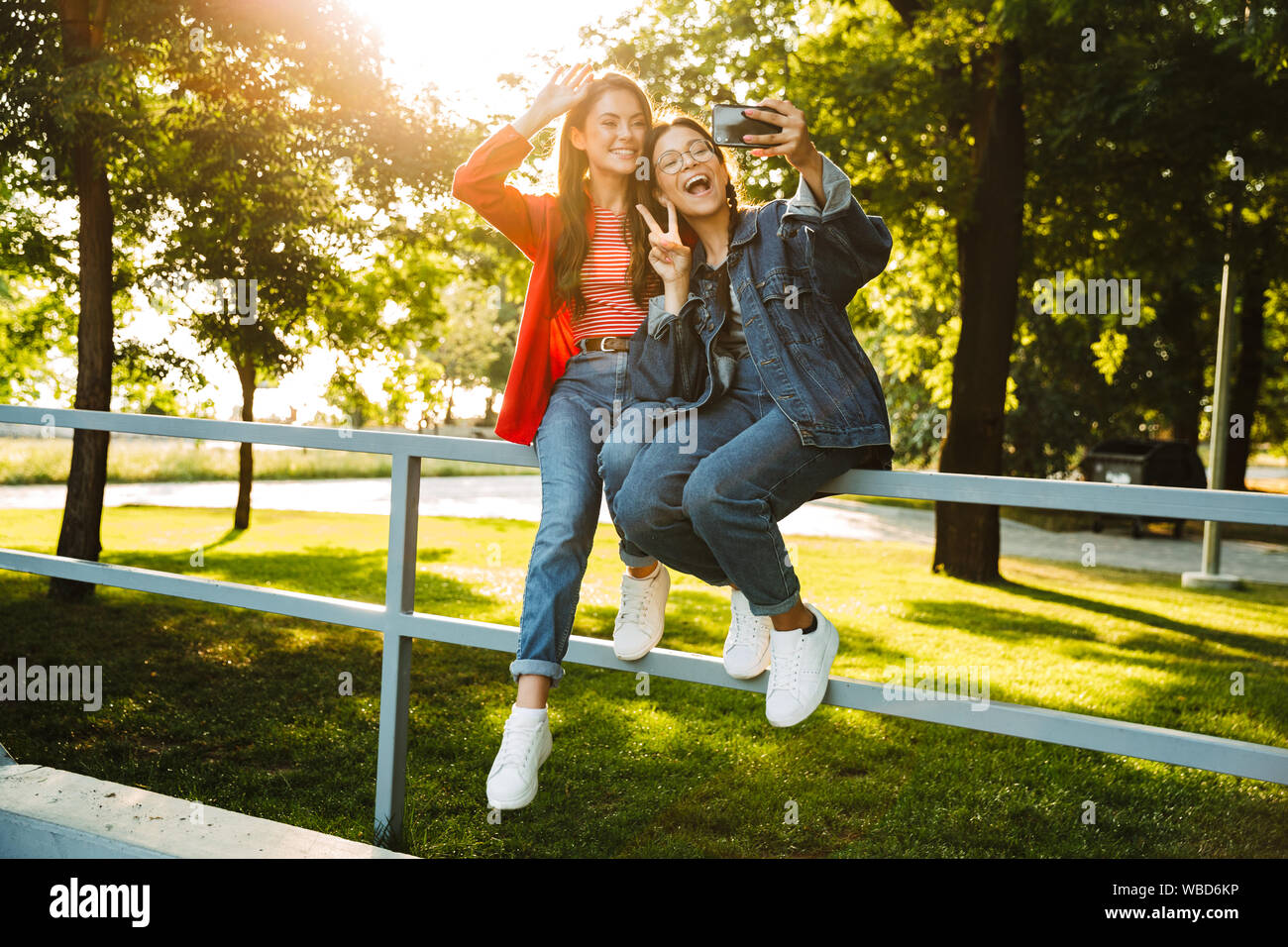 Image de deux élèves filles heureux de prendre photo sur téléphone portable et selfies gesturing while sitting on chanter la paix railing in green park Banque D'Images