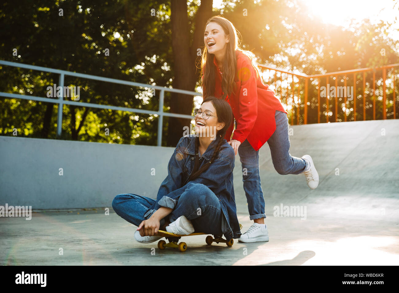 Image de deux filles de joie vêtu de vêtements en denim de rire et équitation skateboard ensemble dans skate park Banque D'Images