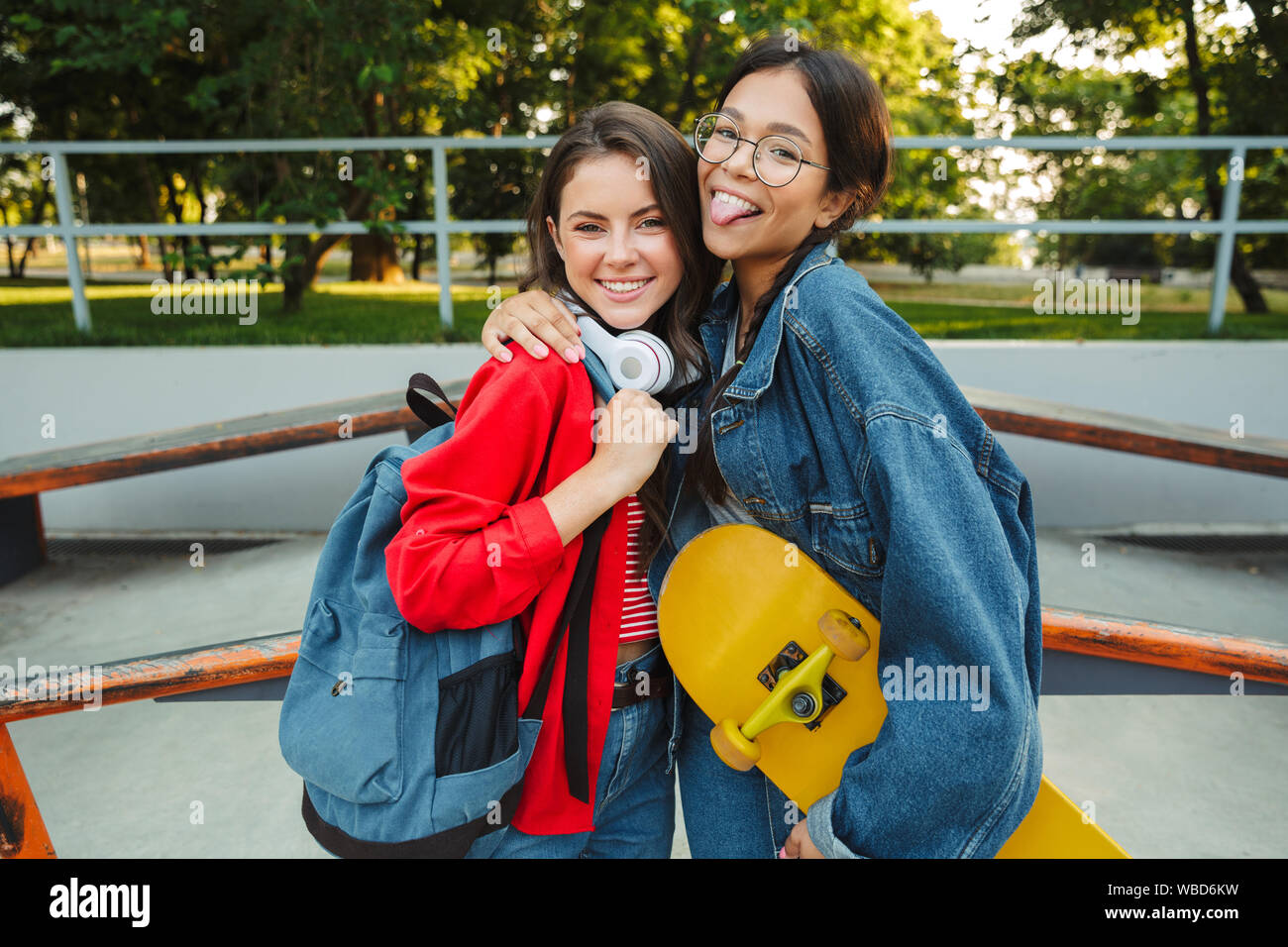Image de deux funny girls habillés en vêtements de denim en souriant et serrant ensemble tout en maintenant en skateboard skate park Banque D'Images