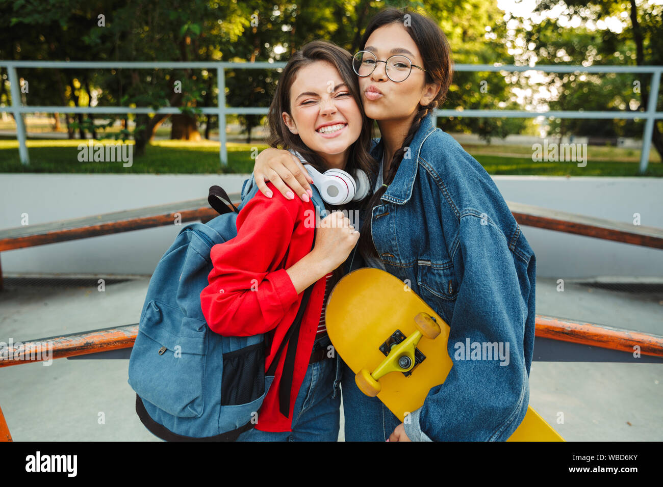 Image de deux jolies filles habillés en vêtements de denim en souriant et serrant ensemble tout en maintenant en skateboard skate park Banque D'Images