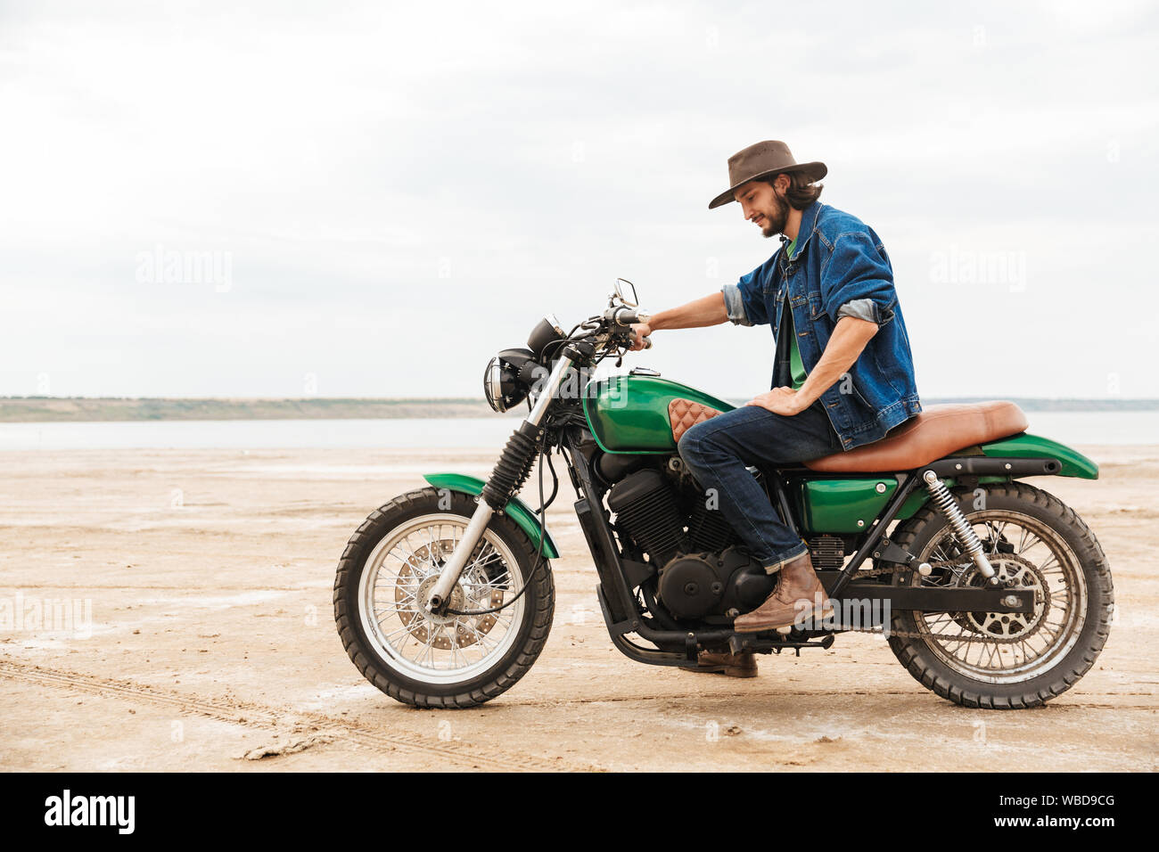 Vue latérale d'un beau jeune homme portant tenue décontractée assis sur une moto à la plage Banque D'Images